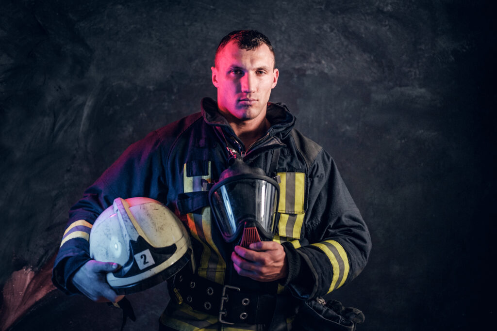 A firefighter in full gear stands against a dark background with red light highlighting part of his stern face, symbolizing strength and resilience in holistic trauma treatment.