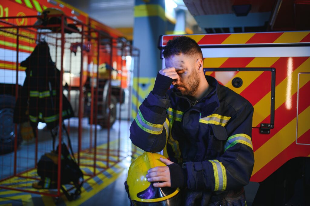 A firefighter in uniform sits on a bench outside the fire station with his head down and his hands covering his face, illustrating the emotional distress and persistent feelings of hopelessness experienced by first responders.






