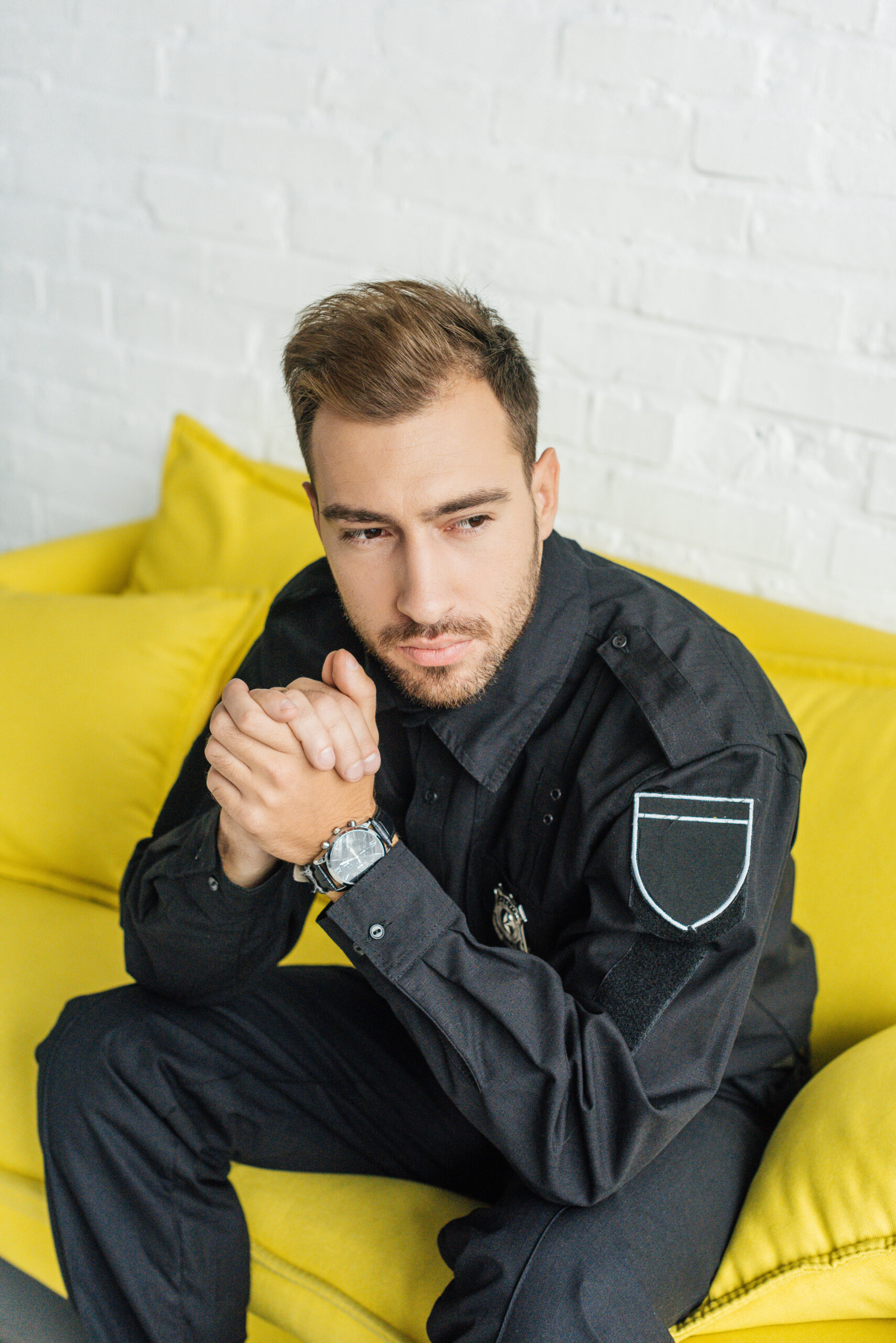 A police officer sitting on a couch with hands clasped, contemplating his mental health, highlighting the psychological symptoms often faced by first responders.