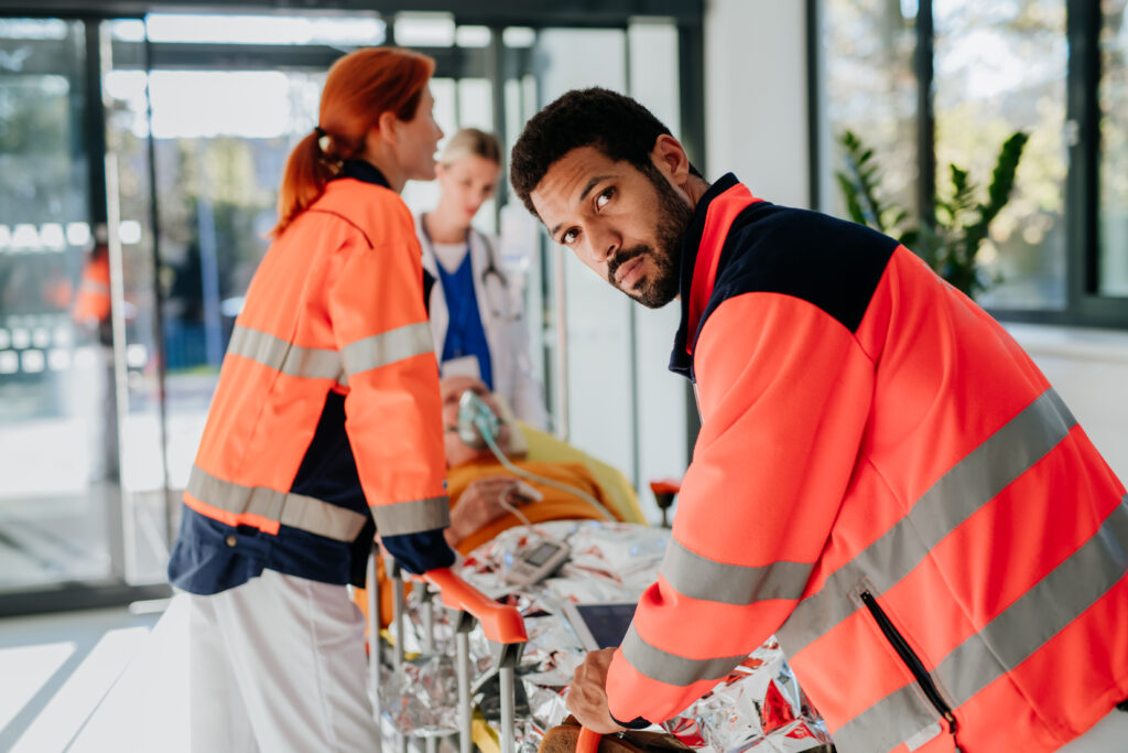 A paramedic pushes a patient on a stretcher into the hospital, looking back over his shoulder with a concerned expression, highlighting the high-stress environment and the potential for substance use as a coping mechanism among first responders.