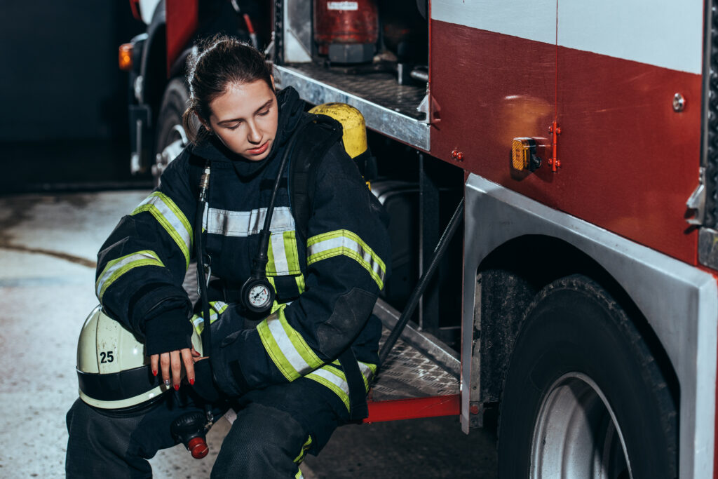 A woman firefighter in uniform stands outside the fire station, looking down with an expression of exhaustion, illustrating the physical symptoms of anxiety and stress faced by first responders.