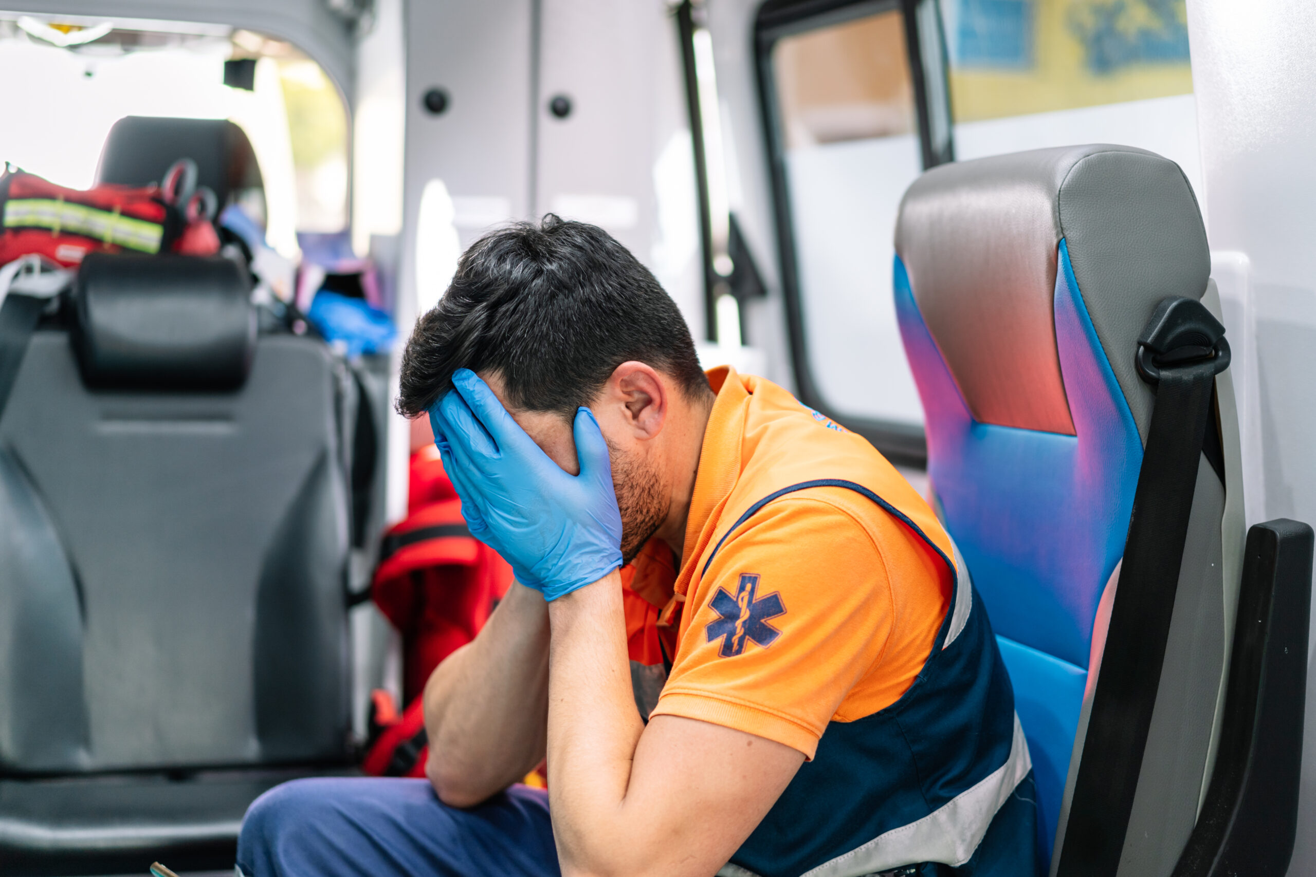 A paramedic in uniform sits on the back of an ambulance with his head in his hands, looking distressed, symbolizing the anxiety and mental health challenges faced by first responders.